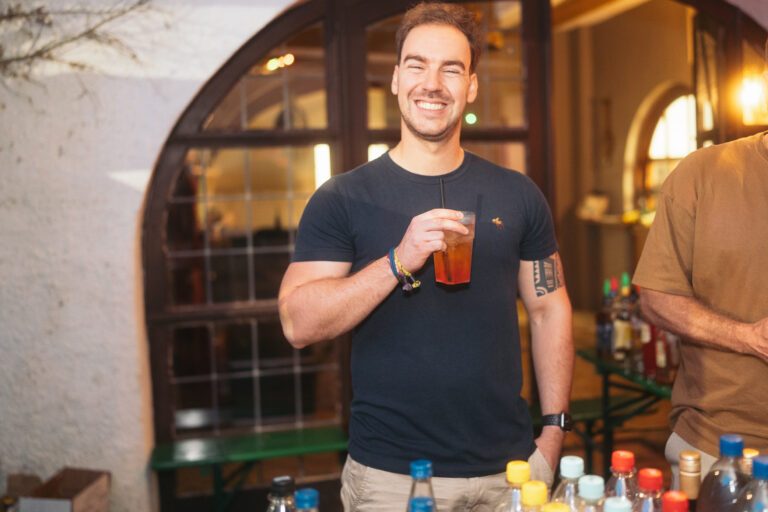 Mann mit dunklem T‑Shirt lächelt und hält ein Getränk in einem Glas, Hintergrund mit Bar und Flaschen, Bezug zu Wild Woods und Trockendock Events