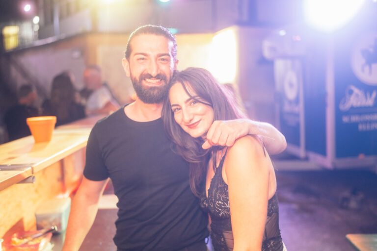 Wild Woods – zwei lächelnde Personen am Barbereich, Teil von Trockendock Events, warme Lichtstimmung mit bunten Lichtern im Hintergrund, Outdoor-Event Atmosphäre.