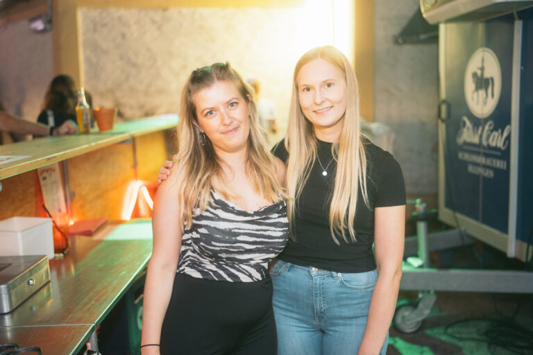 Zwei freundliche Frauen stehen im Barbereich des Wild Woods Events von Wild Woods und Trockendock Events, warme Farbtöne, Neonlicht im Hintergrund, freundliches Lächeln.