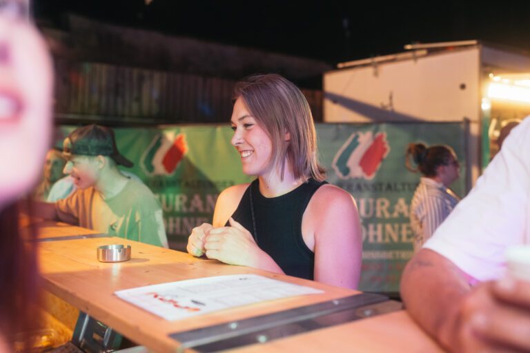 Eine junge Frau mit kurzem lilafarbenem Haar und schwarzem Top lächelt an einem Holztresen bei einer nächtlichen Wild Woods-Atmosphäre von Trockendock Events; im Hintergrund Banner mit SonnenDeck-Logo und diffusem Licht.