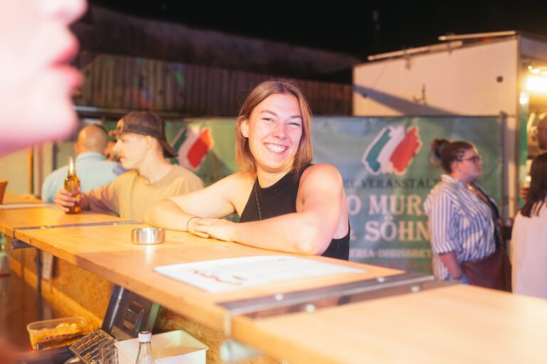Lächelnde Frau am Barbereich von Wild Woods, umgeben von Ambiente mit bunten Lichtakzenten, Banner im Hintergrund, handsom bar und entspannt wirkend