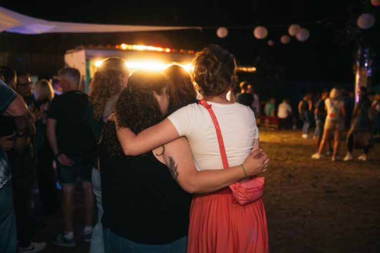 Zwei Freundinnen umarmen sich lächelnd bei einer Nachtveranstaltung von Wild Woods, organisiert von Trockendock Events; im Hintergrund bunte Lichter, eine Festivalatmosphäre, eine rote Umhängetasche und warme Beleuchtung prägen die Szene.