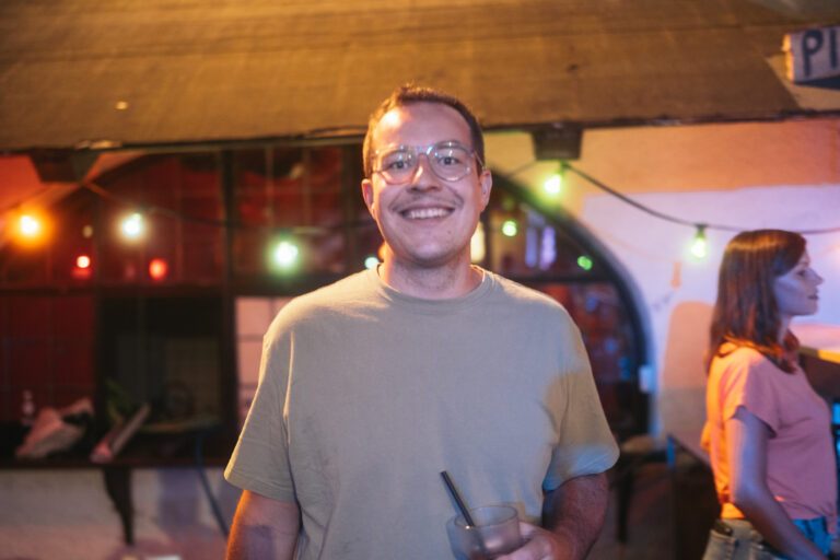 Wild Woods Gast bei Trockendock Events, lächelnd, Brille, beiges Shirt, Trinkglas mit Strohhalm; bunte Neonlichter im Hintergrund schaffen eine lebendige Nachtstimmung