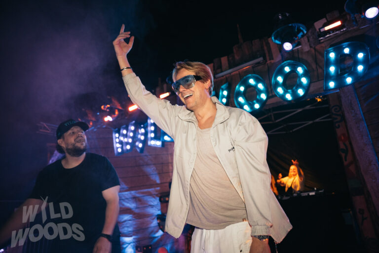 Beim Wild Woods Festival hebt ein DJ mit Sonnenbrille die Hand, während blaue Lichtshow und Nebel über der Bühne strahlen.