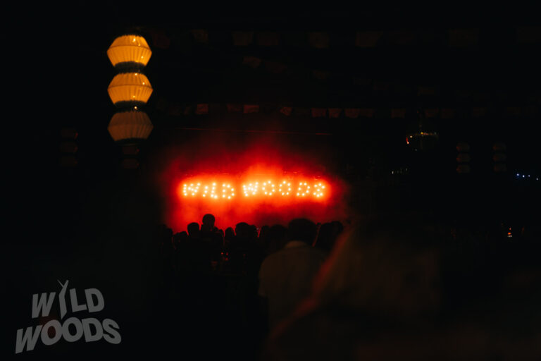 Stimmungsvoller Moment beim Wild Woods Festival: leuchtende Bühne mit roten und orangefarbenen Lichtstrahlen sowie „Wild Woods“ als Show-Text