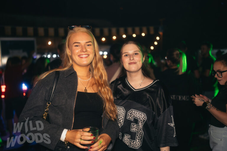 Zwei Besucherinnen bei Wild Woods Festival im Märchenwald, lachend vor grüner Lichtstimmung und Bühnenlichtern in der Nacht