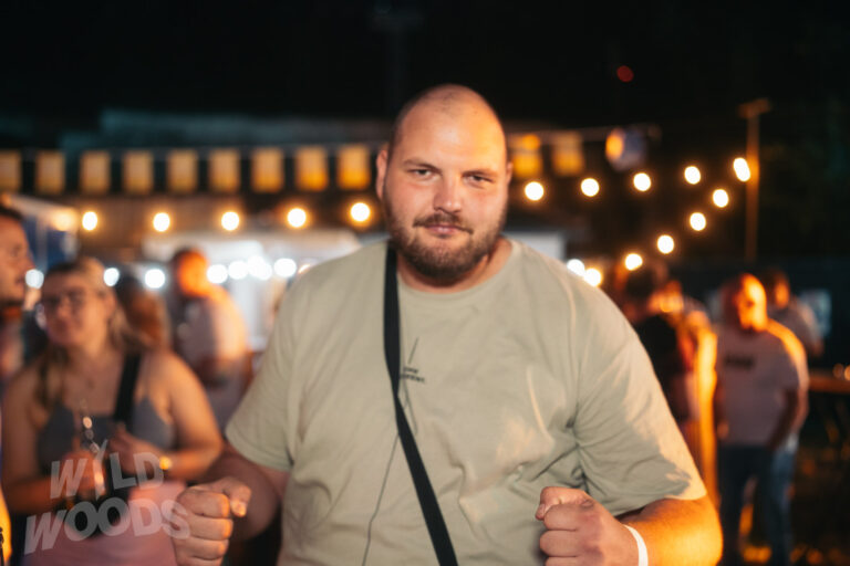 Beim Wild Woods Festival steht ein Festivalgast mit Bart im warmen, orangefarbenen Licht vor einer Reihe leuchtender Lichter und unscharfen Kulissen.