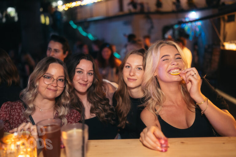 Freundinnen beim Wild Woods Festival im Märchenwald, umgeben von warmen goldenen Lichtern, mit Getränken und lebhafter Stimmung