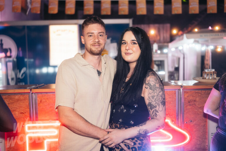 Bei Wild Woods Festival posieren zwei Besucher vor einer Bühne mit orangefarbenem Neonlicht, im Hintergrund warme Lichtketten und Bar-Atmosphäre.