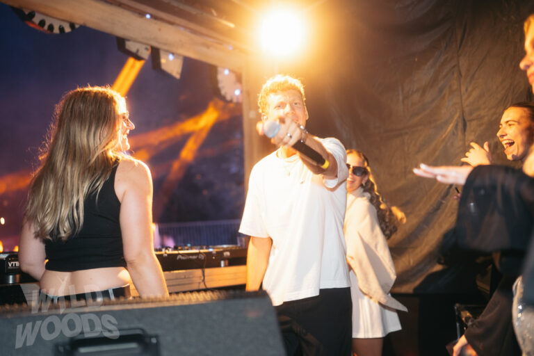 Stimmungsvolle Szene beim Wild Woods Festival: ein DJ mit Mikrofon auf der Bühne, umgeben von Publikum im warmen Licht und im Märchenwald mit elektronischen Beats.