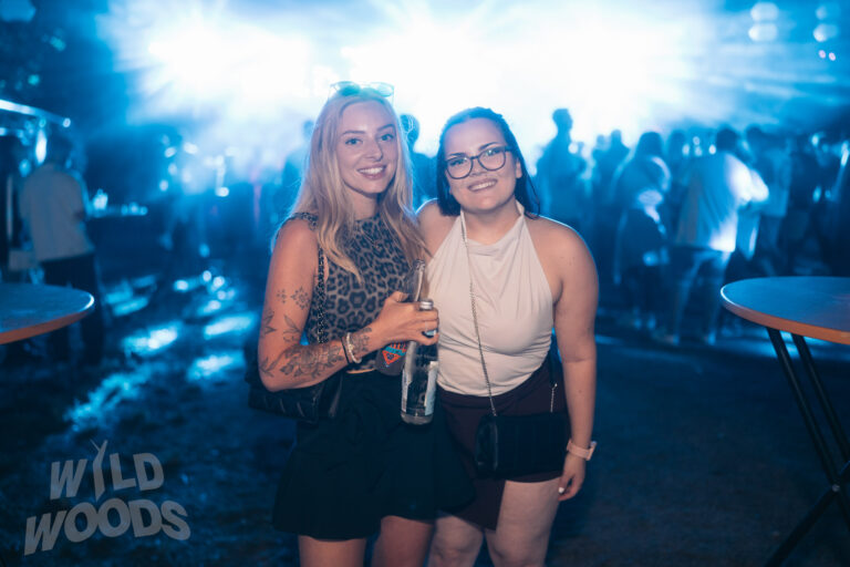 Beim Wild Woods Festival 2025 stehen zwei Besucherinnen blau beleuchtet und lächelnd zusammen, eine mit Leopardenmuster, im Hintergrund eine volle Tanzfläche.