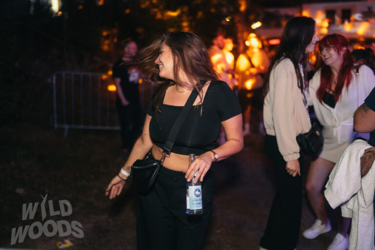 Beim Wild Woods Festival genießen Besucher den Abend bei warmem Licht und nächtlicher Stimmung, eine Person mit schwarzem Outfit und Getränk in der Hand.