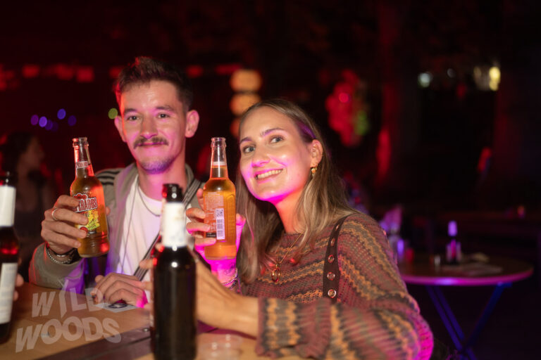 Wild Woods Festival: zwei Besucher mit „Wild Woods“-Aufdruck, halten Flaschen im magenta-lila Licht bei elektronischer Party im Märchenwald.